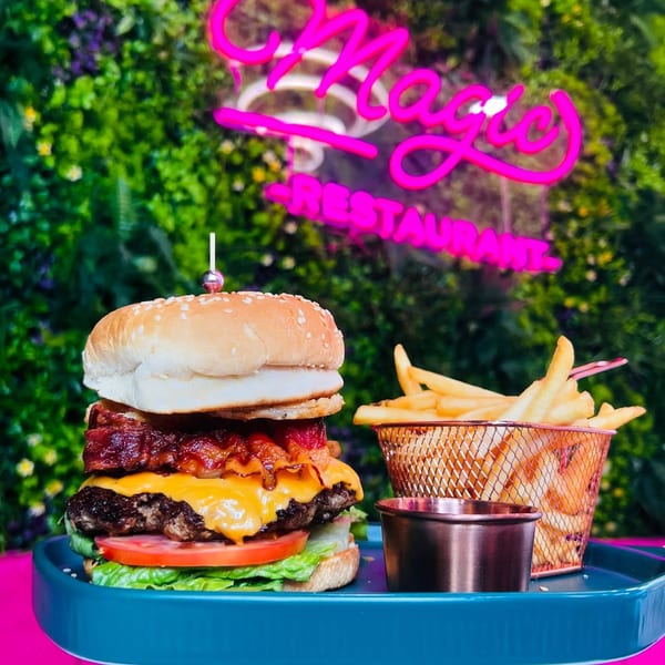 Magic Restaurant's bacon cheeseburger with fries and sauce on a blue tray, against a green wall with a neon sign.