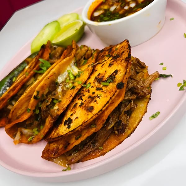 Close-up of four birria tacos with limes, grilled jalapeño, and a side of consommé.