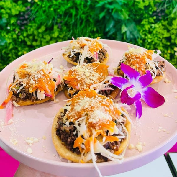 Five Latin-American sopes topped with meat, cabbage, sauce, cheese, and a purple orchid on a pink plate.